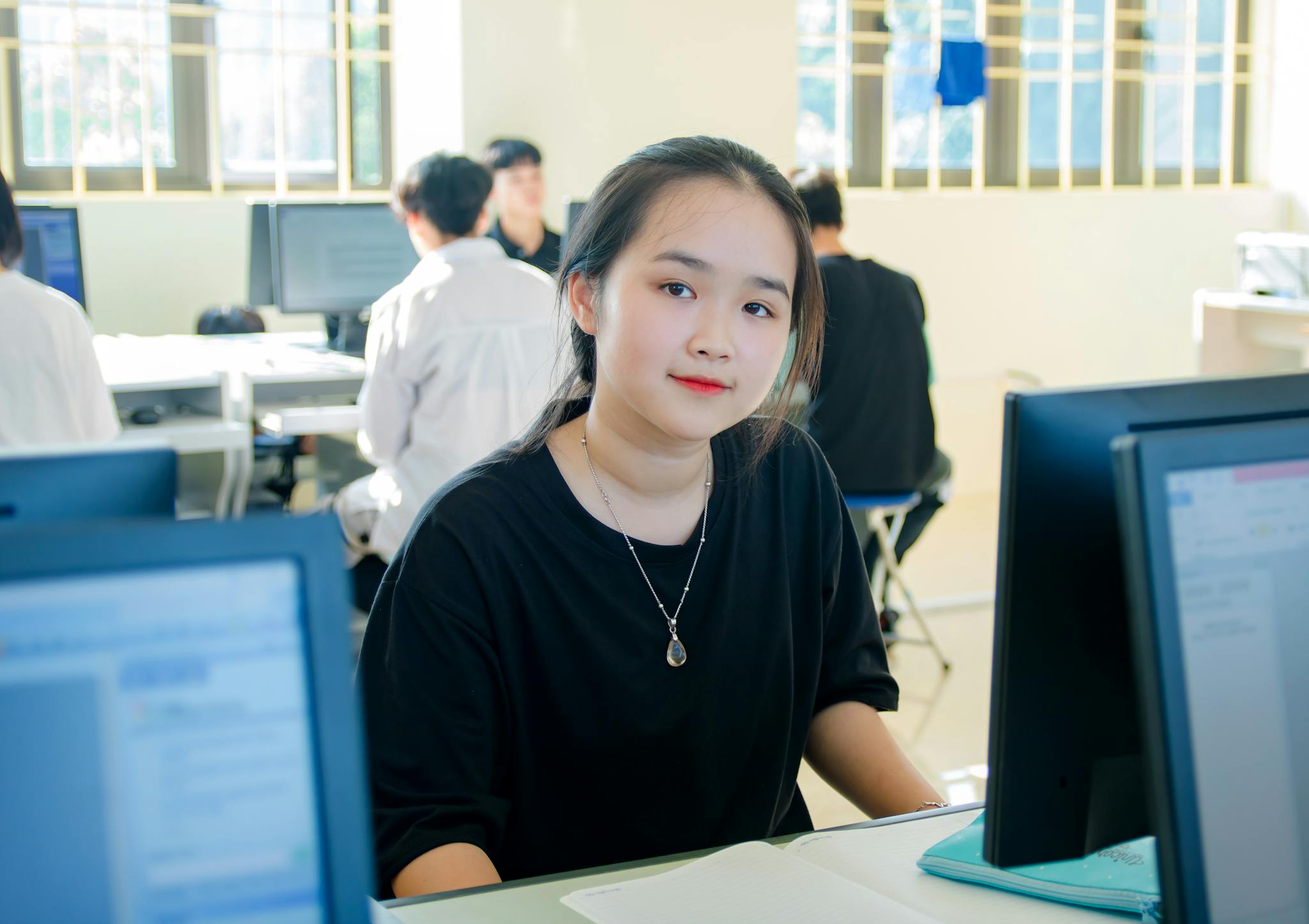 A young woman sits at a desk in a computer lab, smiling into the camera.